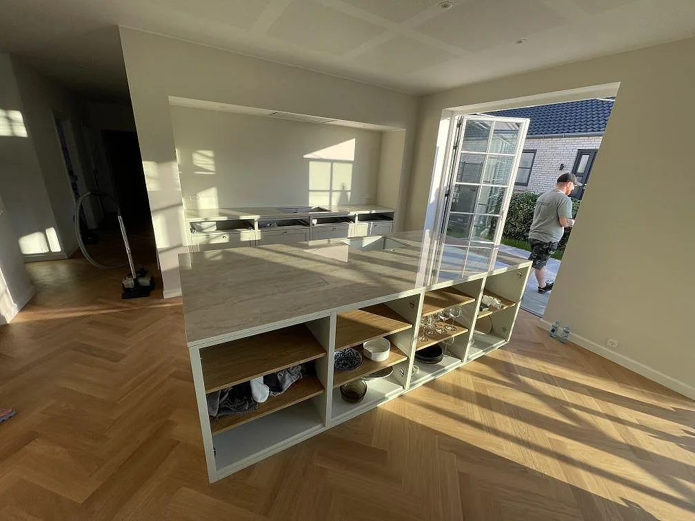 Helhetsbild av kök i Kastrup med stor köksö, polerad Dekton Arga, fiskbensgolv och vita stommar under montering.