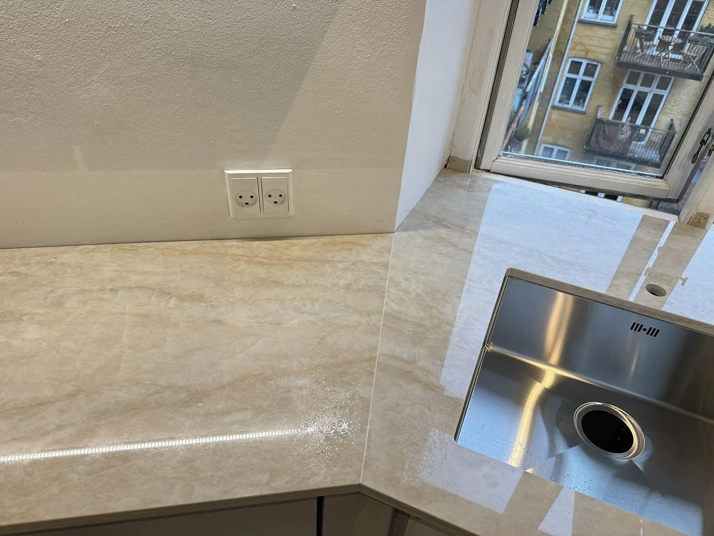 Köksbänkskiva i polerad Dekton Arga, ljus keramik med rostfri diskho och fönster i ett L-format kök i København N.