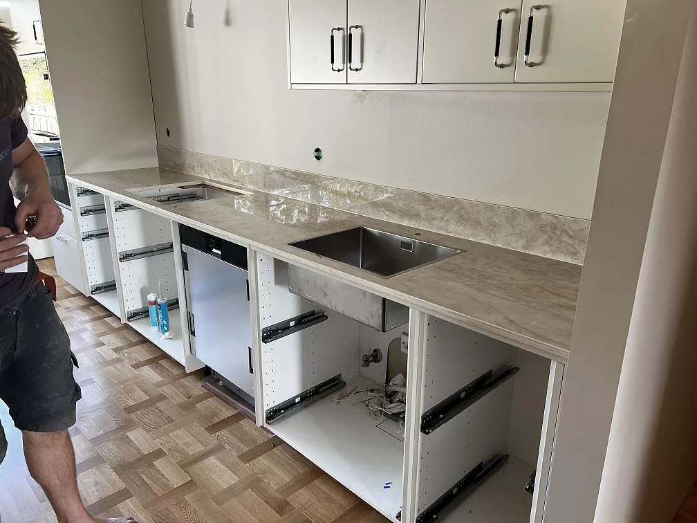 Lång bänkrad i polerad Dekton Arga keramik med stänkskydd, rostfri diskho och vita köksstommar under montering i Lund.