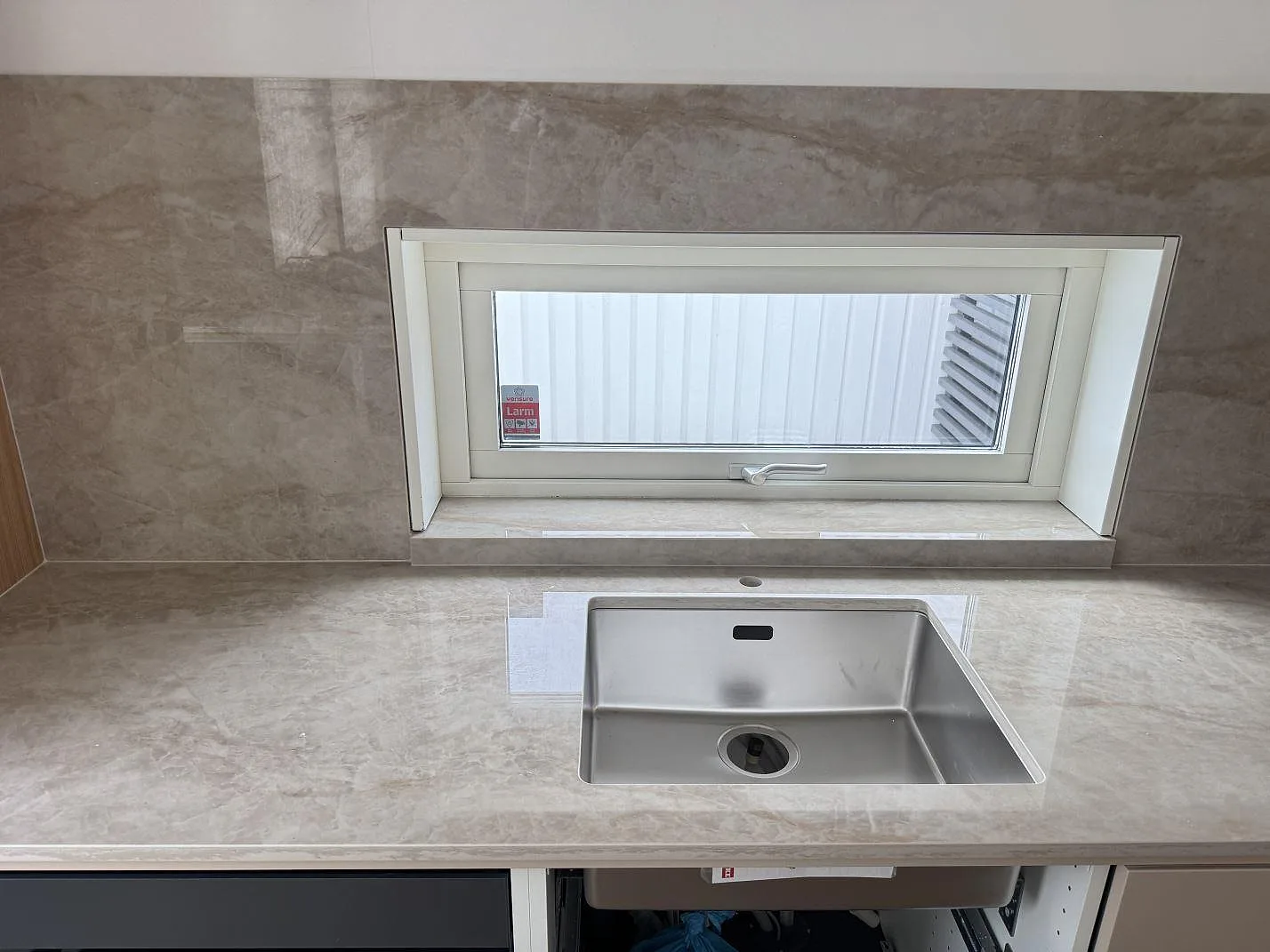 Köksbänkskiva i ljus krämbeige Dekton Arga, polerad, med underlimmad rostfri diskho och heltäckande stänkvägg i samma keramik vid fönstret i ett kök i Malmö.