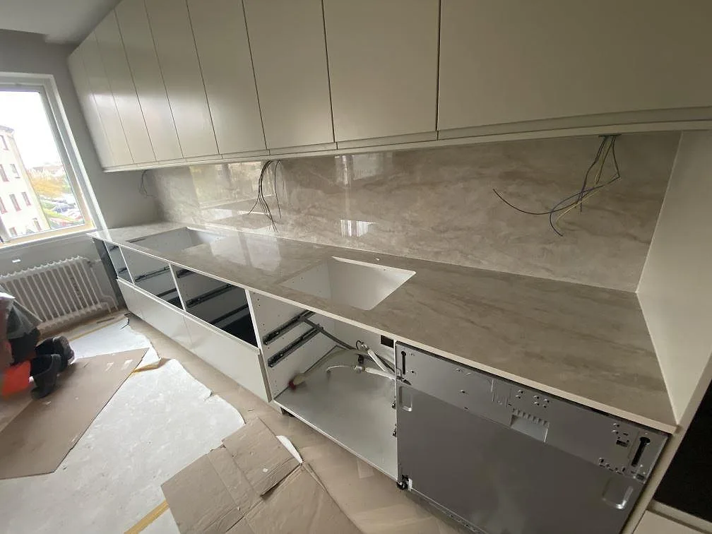 Köksbänkskiva i cremebeige Dekton Arga, polerad, med heltäckande stänkskydd och underlimmad diskho i ett kök under montering i Mölndal.