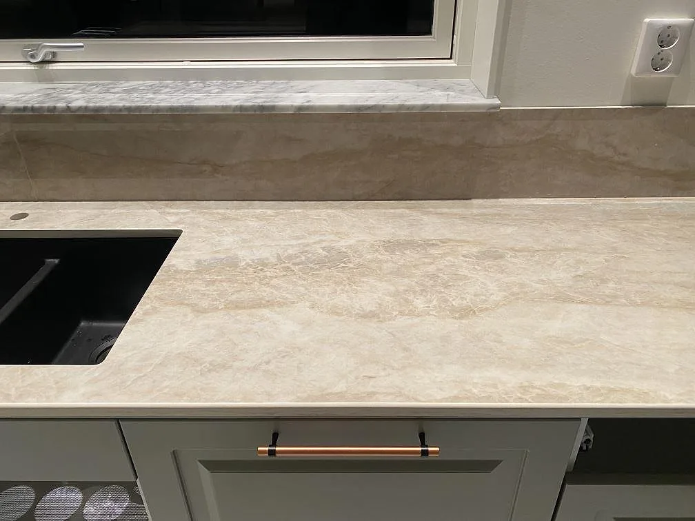 Köksbänkskiva i Dekton Arga, polerad keramik i krämbeige marmorlook, med underlimmad svart kompositdiskho och uppvikt stänkkant i ett kök i Nybro.