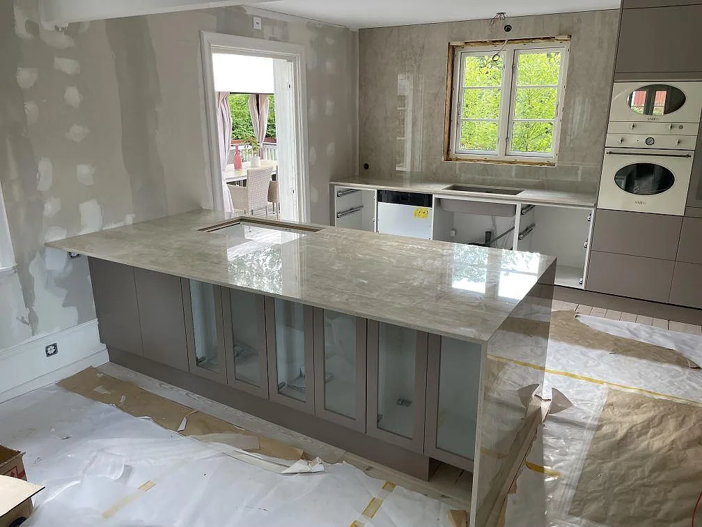 Polerad köksbänkskiva i Dekton Arga keramik med köksö, neddragen sidopanel, diskho och väggbeklädnad i ett kök i Sandviken.