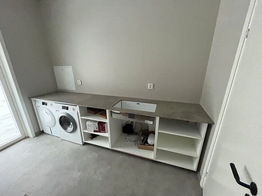 Tvättstugebänk i polerad Dekton Arga med tvättmaskin och ho i en separat tvättstuga under montering i Sigtuna.
