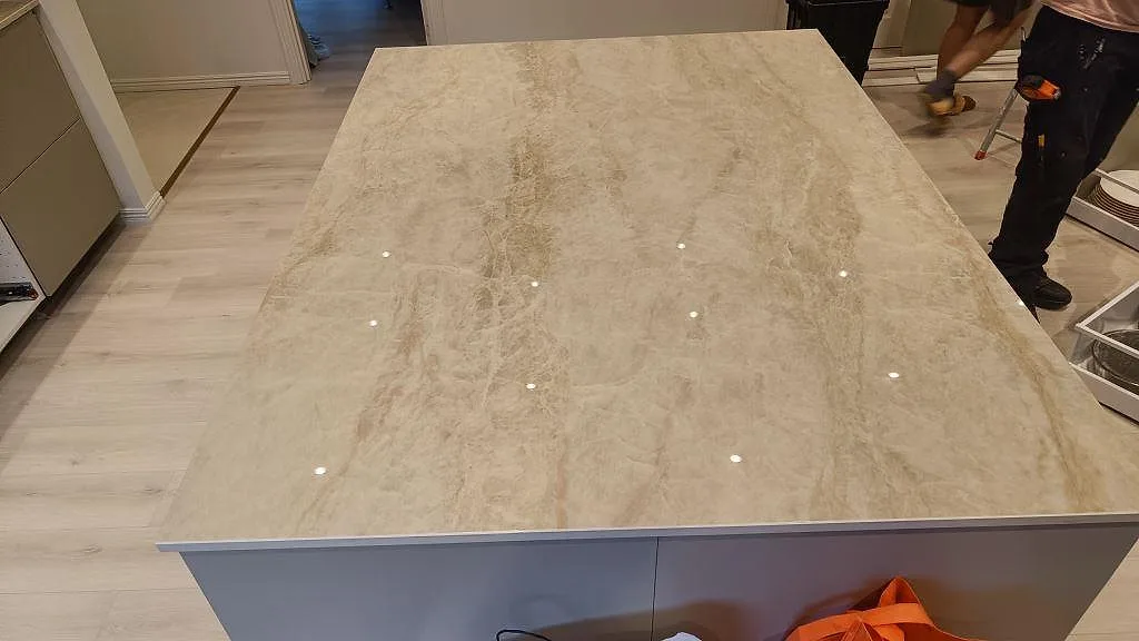 Ytnära vy av den stora köksön i polerad Dekton Arga med beige ådring och öppna stommar under montering i Skövde.