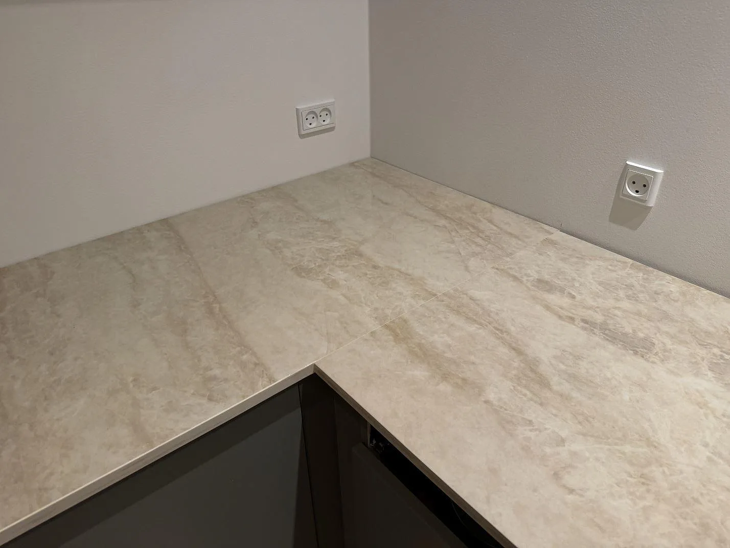Köksbänkskiva i krämigt beige Dekton Arga, polerad keramik, i L-format längs två väggar med synlig ådring och skarv i ett kök i Skovlunde.