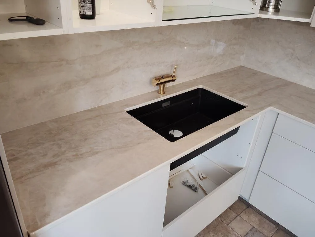 Köksbänkskiva i polerad Dekton Arga med matchande stänkskydd, svart underlimmad diskho och mässingsfärgad blandare i ett kök i Solna.