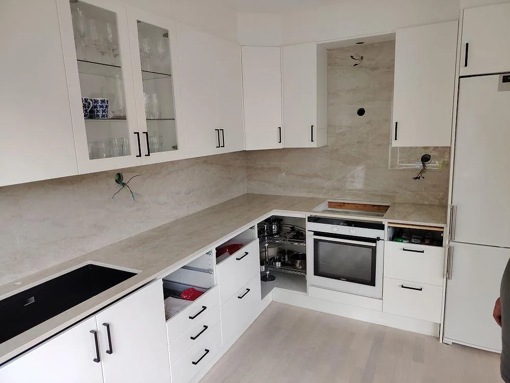 Polerad keramisk köksbänkskiva och väggbeklädnad i Dekton Arga i ett vitt vinkelkök med inbyggnadsugn och hällzon i Solna.