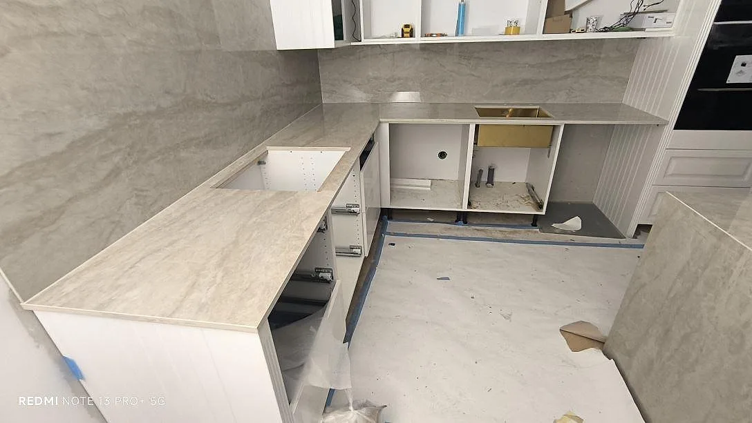 L-formad köksbänkskiva i polerad Dekton Arga med ljus ådring, väggbeklädnad och öppna köksstommar under montering i Spånga.