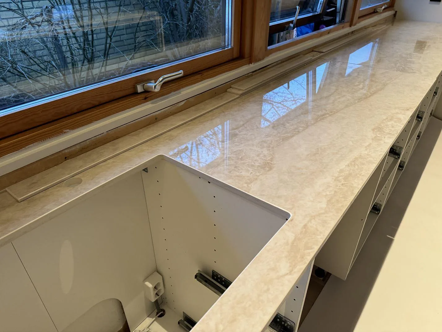 Lång köksbänkskiva i polerad Dekton Arga vid träfönster med öppning vid diskbänksdelen i Staffanstorp.