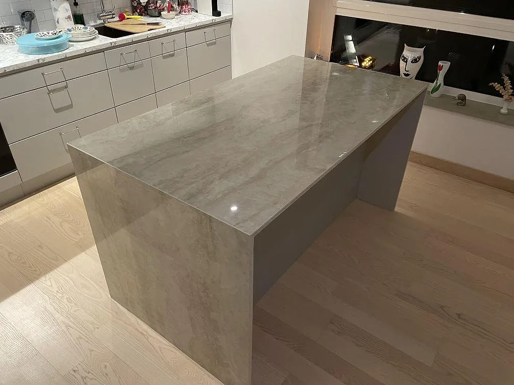 Fristående köksö i cremebeige Dekton Arga, 20 mm polerad, med nedvikt stensida mot trägolv och en ljus marmorlik bänkrad i bakgrunden i ett kök i Västervik.