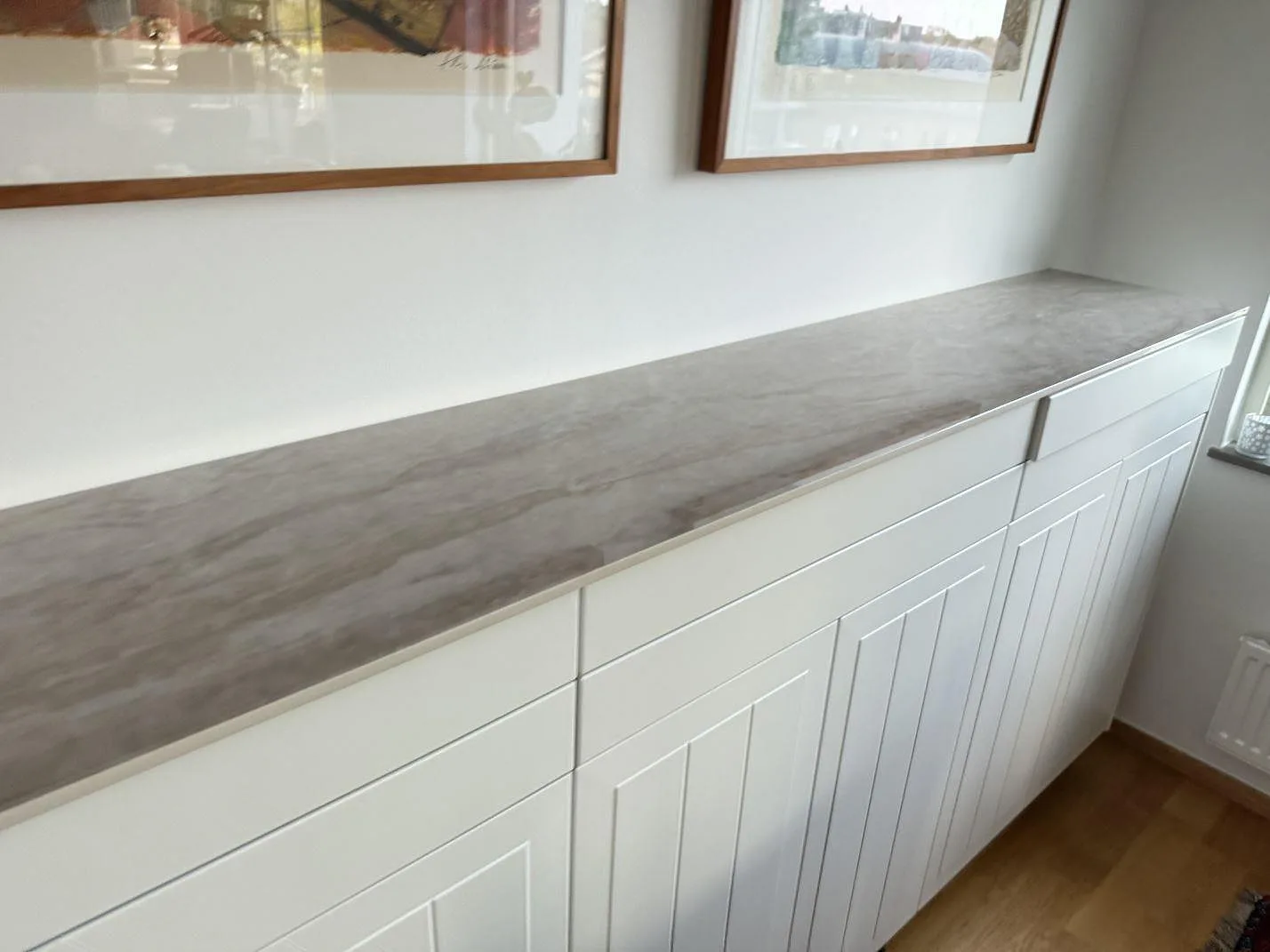 Lång bänkskiva i polerad Dekton Arga keramik på vit förvaringsrad i Vellinge, med gråbrun ådring under tavlor.