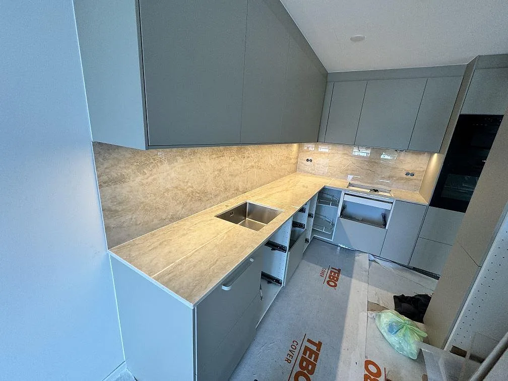Översiktsbild av kök i Ystad med polerad Dekton Arga som L-formad köksbänkskiva och stänkskydd under överskåpen.