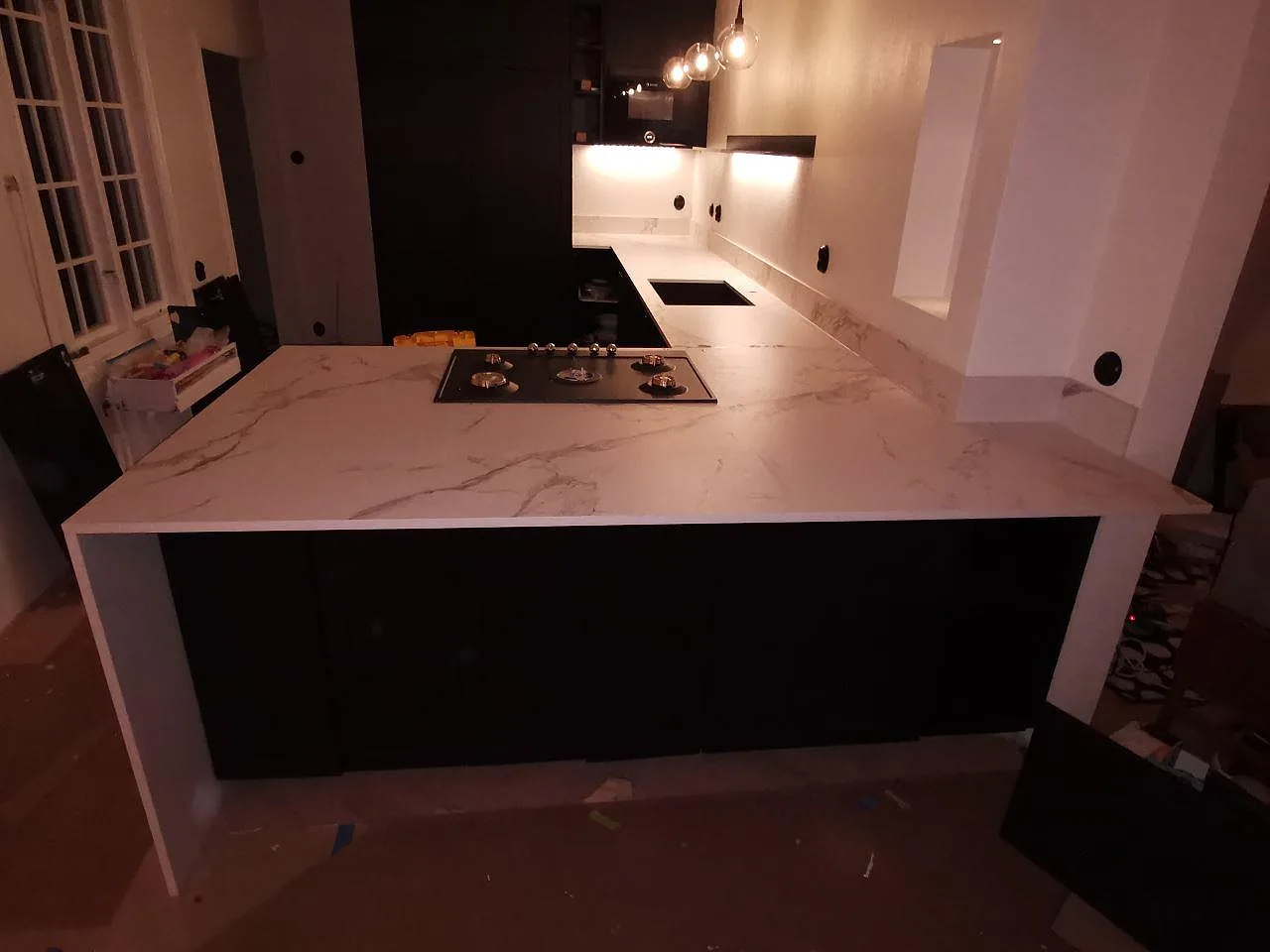 Köksbänkskiva i vit Dekton Aura med marmorlik ådring på en halvö med infälld häll, mot mörka luckor i ett kök i Varberg.