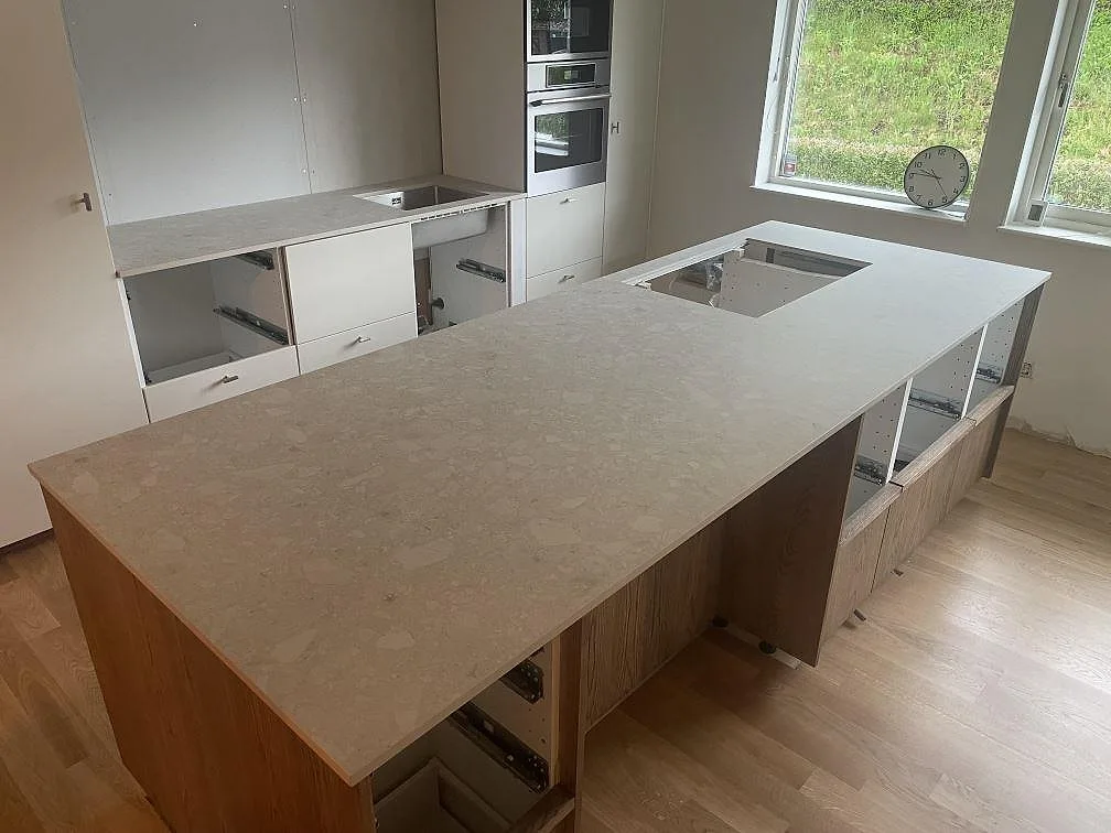Köksbänkskiva och stor köksö i matt Dekton Ava, varmt cremefärgad keramik med terrazzomönster, med ekfronter och infälld induktionshäll i ett kök under montering i Falun.