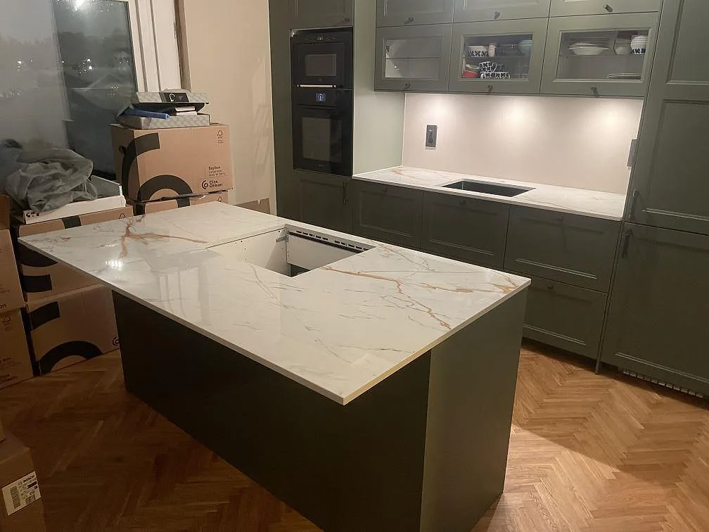 Köksbänkskiva och köksö i vit Dekton Awake med gyllene ådring, polerad, i ett salviagrönt kök i Enskede med underlimmad rostfri diskho och urtag för häll i ön.
