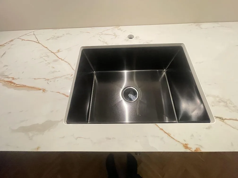 Underlimmad rostfri diskho infälld i polerad Dekton Awake med varm gyllene ådring i ett kök i Enskede, sedd uppifrån.