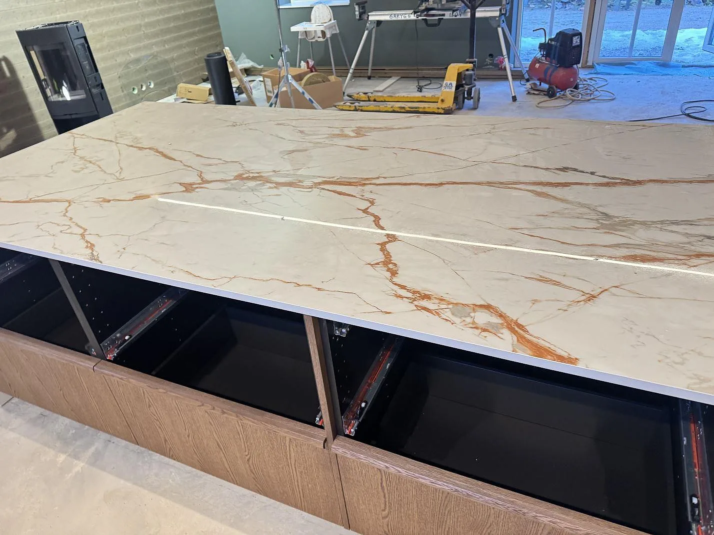 Närbild på lång köksö i polerad Dekton Awake, ljus keramik med grå och rostfärgad ådring, i ett kök under montering i Osby.