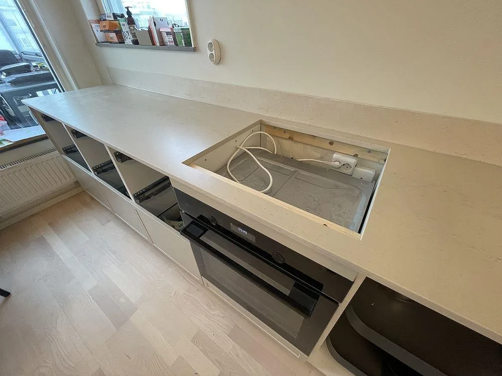 Bänkskiva i matt Dekton Danae med hällöppning framför ugnen och låg stänkkant längs fönsterväggen i Hägersten.