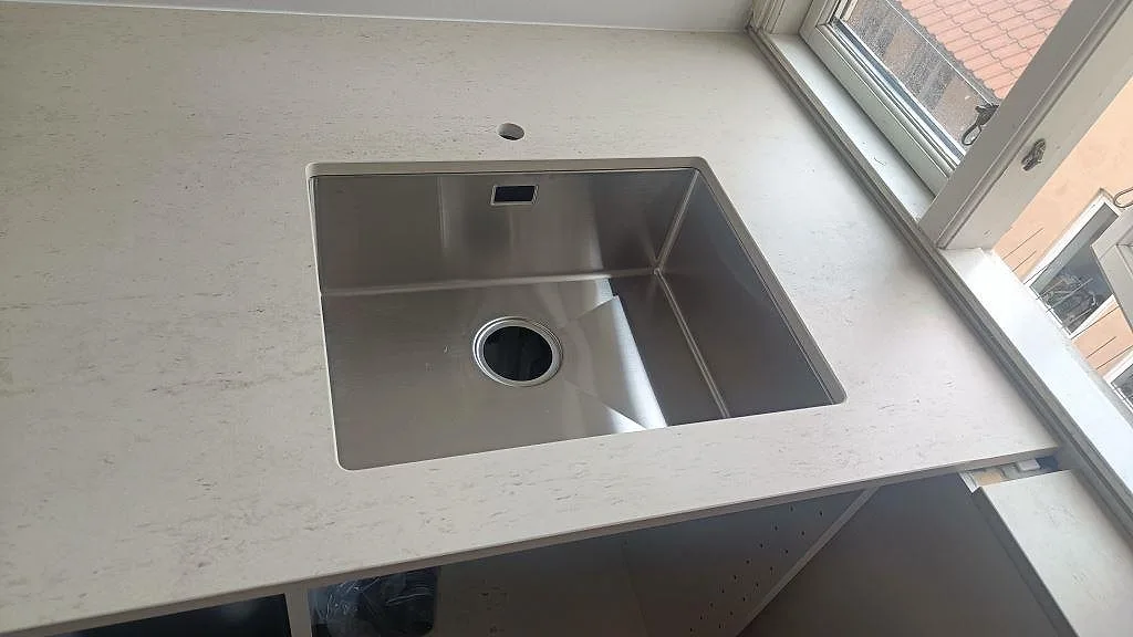Underlimmad rostfri diskho i matt Dekton Danae keramik, monterad vid fönstret i en ljus köksbänkskiva i København N.