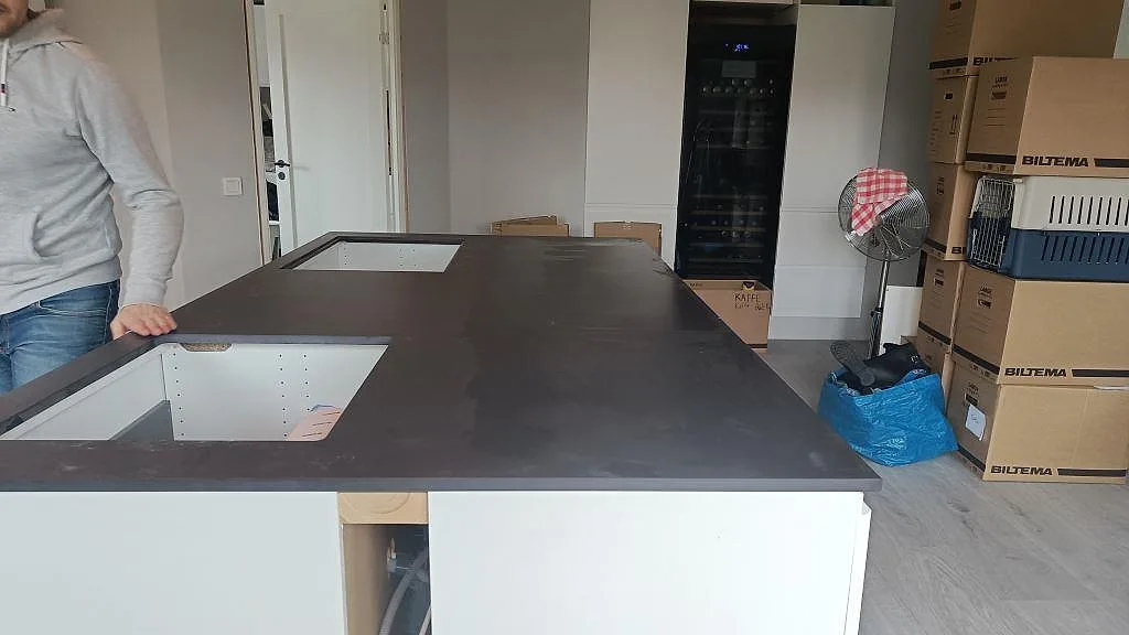 Köksbänkskiva i mörkt grå Dekton Domoos med betonglook på en stor köksö, med två förberedda urtag för underlimmade hoar, i ett kök under montering i Häljarp.