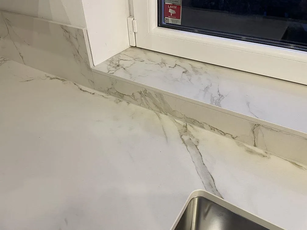 Köksbänkskiva i vit Dekton Entzo, matt keramik med grå Calacatta-lik ådring, som löper fram till fönstret med stenklädd fönsterbänk i ett kök i Ekeby.