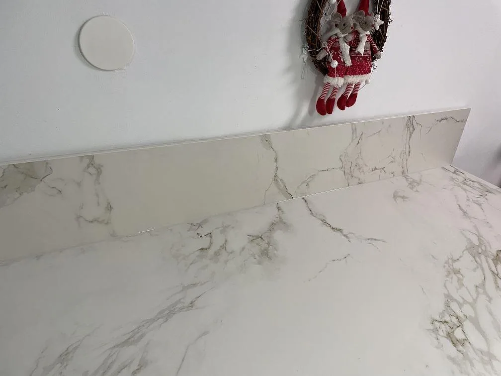 Uppvikt stänkskydd i matt vit Dekton Entzo keramik mot köksbänkskivan, med synlig grå ådring, i ett kök i Ekeby.