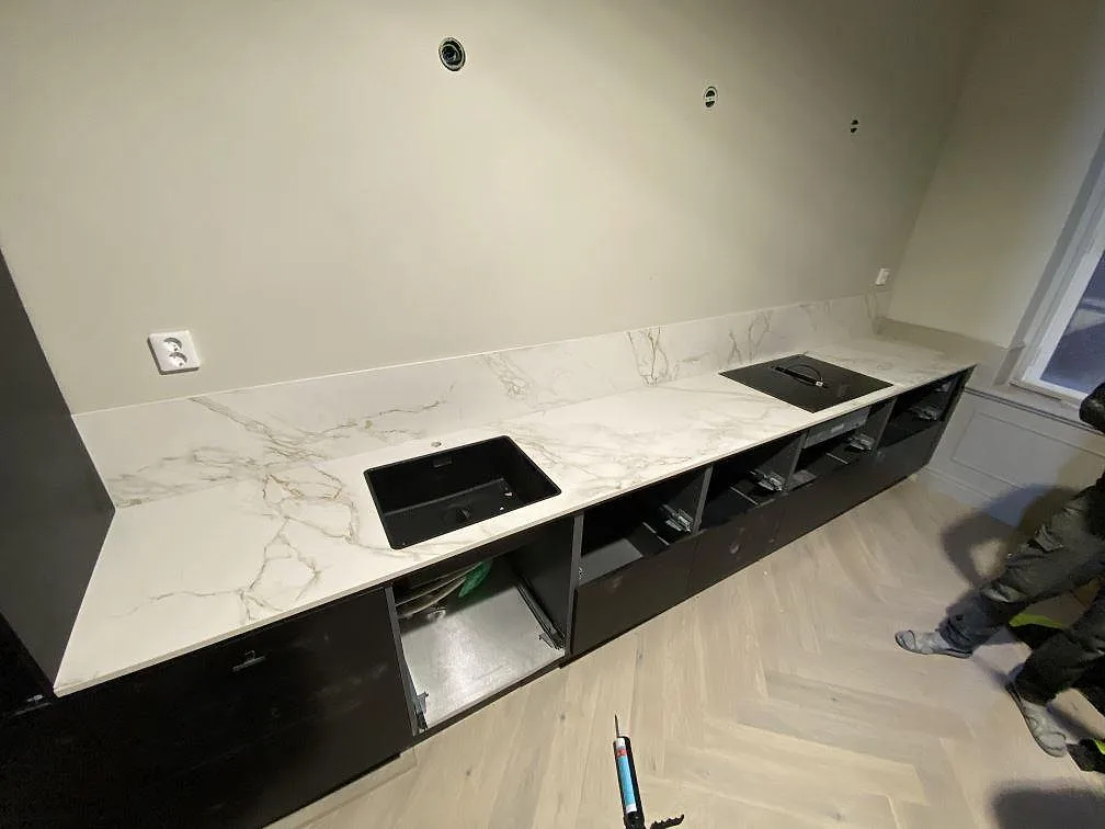 Köksbänkskiva i vit Dekton Entzo med grågyllene ådring, matt yta, längs hela köksraden med underlimmad svart kompositdiskho och plats för häll i ett kök i Göteborg.