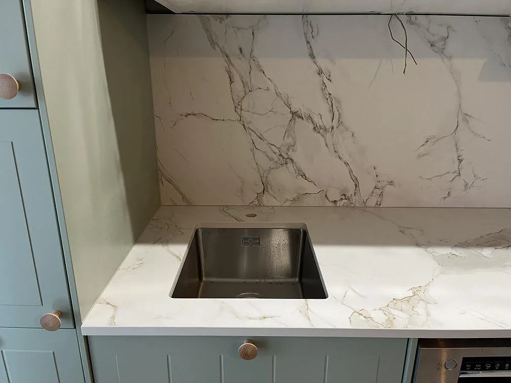 Närbild på köksbänkskivan i matt Dekton Entzo i Hägersten med underlimmad rostfri diskho, kopparfärgade knoppar och heltäckande stänkvägg i vit keramik med grå ådring.