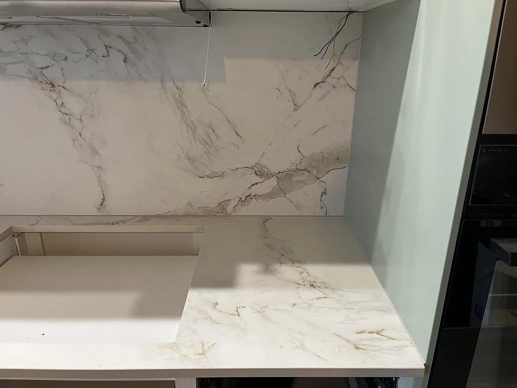 Köksbänkskiva i matt Dekton Entzo i Hägersten med L-vänd bänk, förberett urtag för häll och heltäckande vit stänkvägg intill en ugnshög.