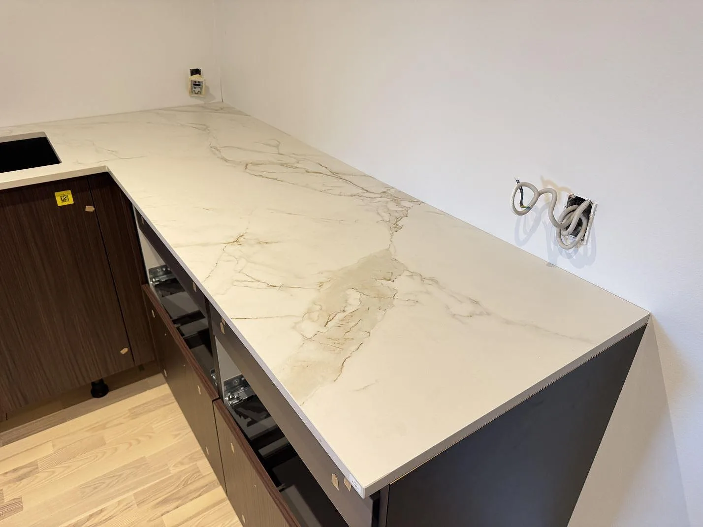 Köksbänkskiva i Dekton Entzo, matt vit keramik med varm guldgrå Calacatta-ådring, längs en L-formad köksrad i ett kök under montering i Hedehusene.