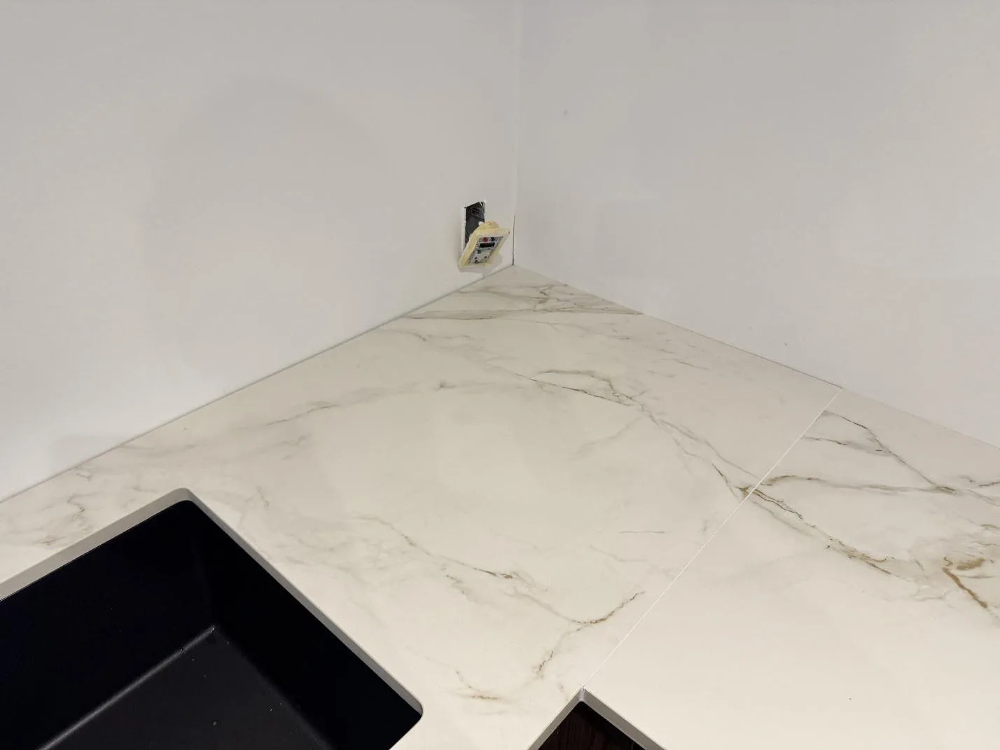 Hörn av köksbänkskiva i vit Dekton Entzo keramik med fin grå ådring och en underlimmad svart kompositdiskho i ett kök i Hedehusene.