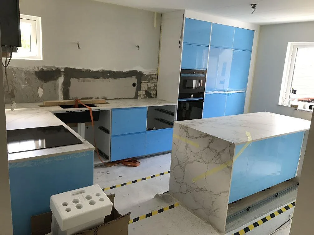 Köksbänkskiva i vit Dekton Entzo, matt, i ett L-format kök i Limhamn med köksö, infälld induktionshäll och blå luckor under montering.