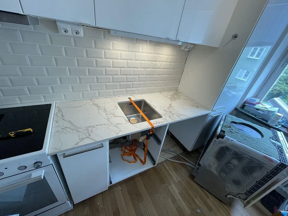 L-format kök i Malmö med matt Dekton Entzo köksbänkskiva, rostfri underlimmad diskho, svart häll och vit kakelvägg under montering.