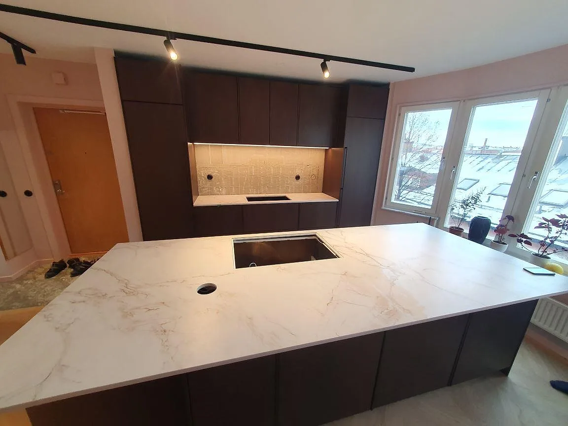 Köksbänkskiva och stor köksö i ådrad vit Dekton Entzo, matt, med urtag för häll i ett ljust kök i Norrköping.