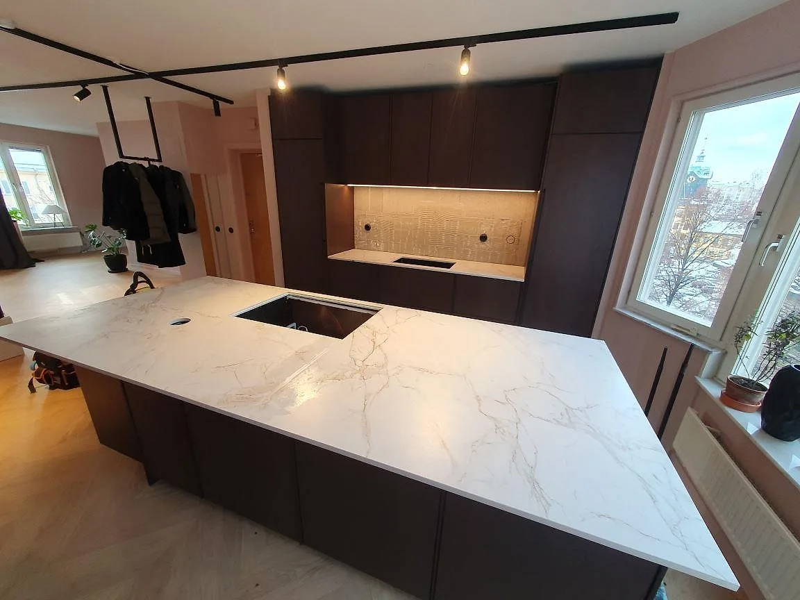 Köksö i vit ådrad Dekton Entzo med urtag för häll mot mörka träluckor i ett kök i Norrköping.