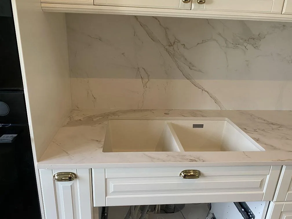 Köksbänkskiva i matt vit Dekton Entzo med underlimmad dubbel komposithoar och heltäckande stänkvägg med grå ådring i ett kök i Svedala.