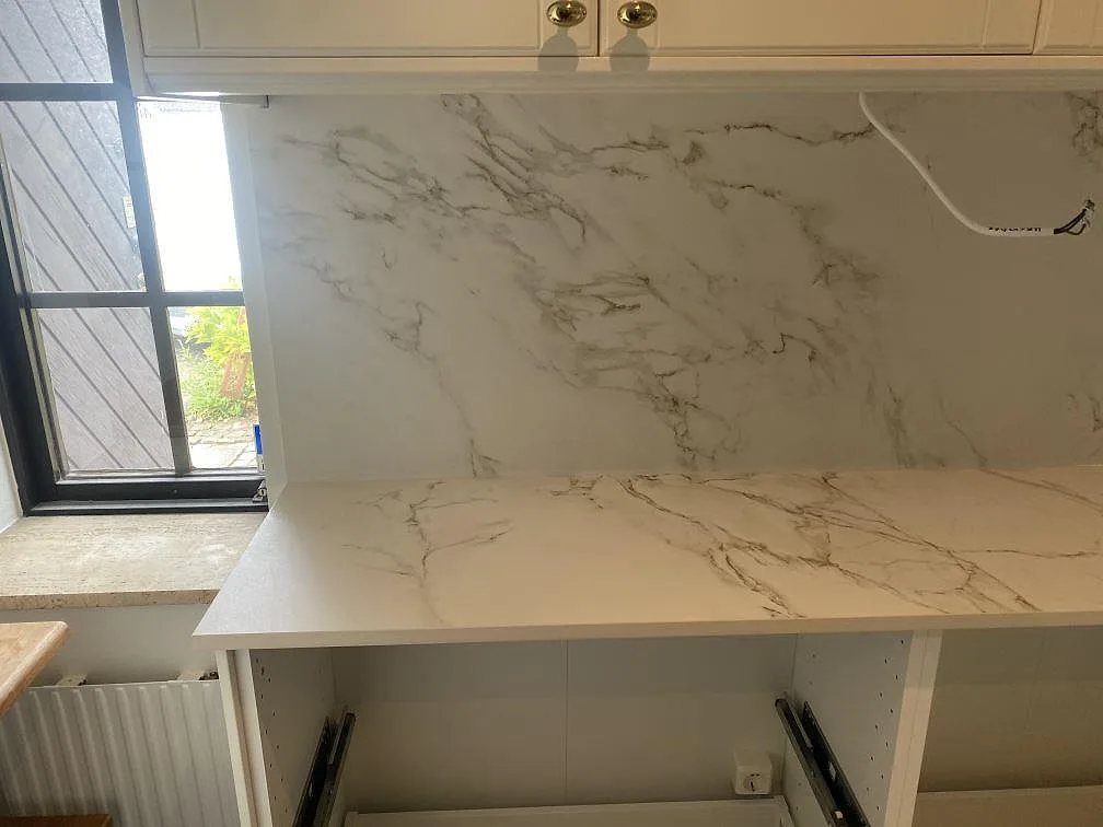 Köksbänk i matt vit Dekton Entzo med markerad Calacatta-ådring och heltäckande stänkvägg vid fönstret i ett kök i Svedala.