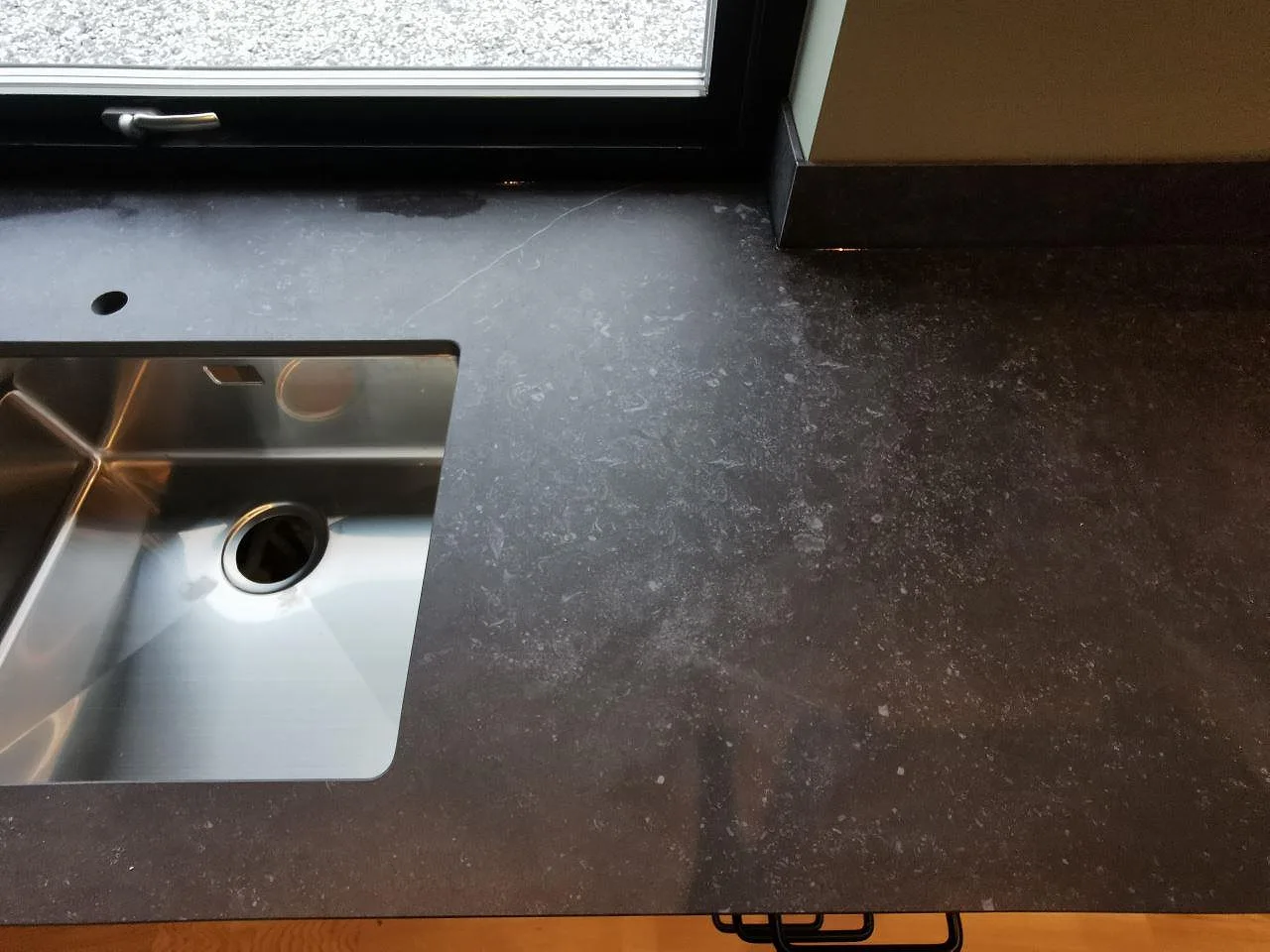 Detalj av bänkskiva i Dekton Fossil vid fönstret med underlimmad rostfri diskho, blandarehål och lågt stänkskydd i samma mörka sten i ett kök i Landvetter.