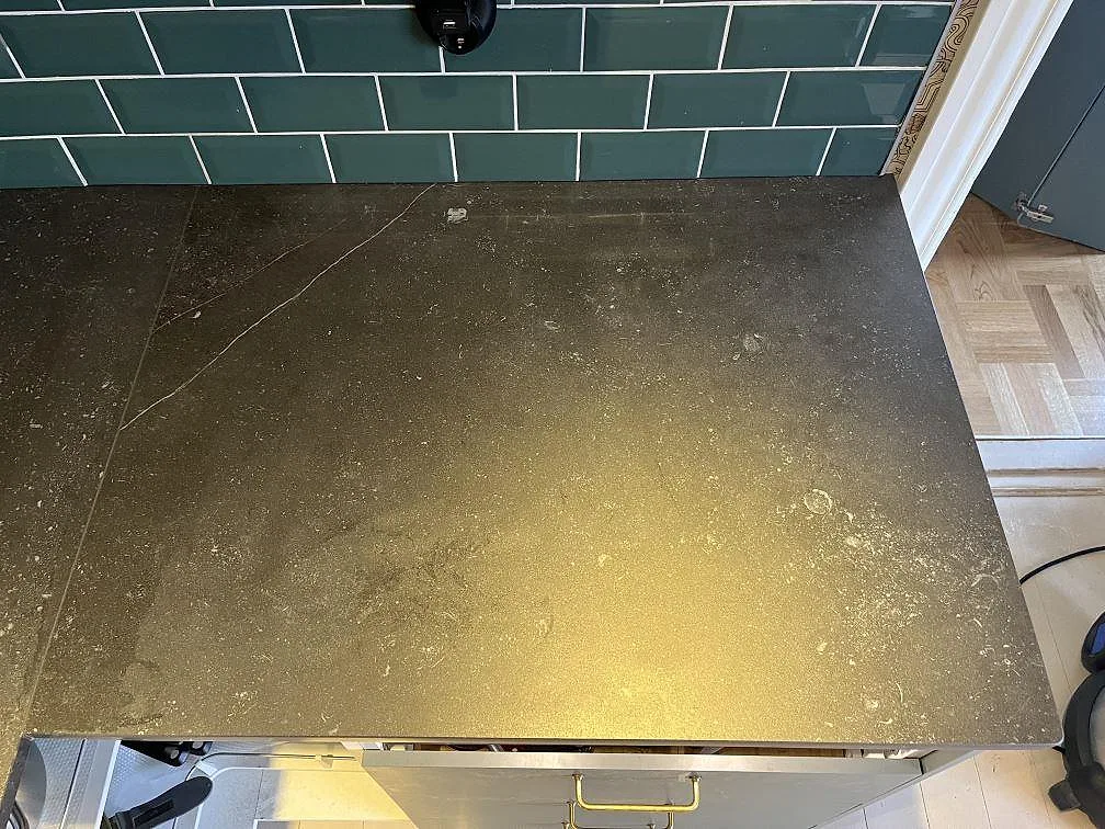 Matt yta på en köksbänkskiva i Dekton Fossil keramik med fossilliknande inslag mot grön kakel i Timmele.