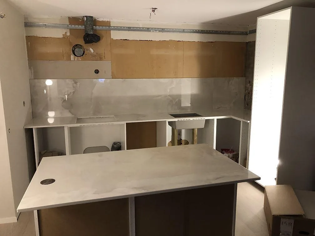 Översikt av ett kök under montering i Lessebo med köksö och bänkrad i Dekton Helena, stänkpaneler i samma sten och urtag för diskho och häll.