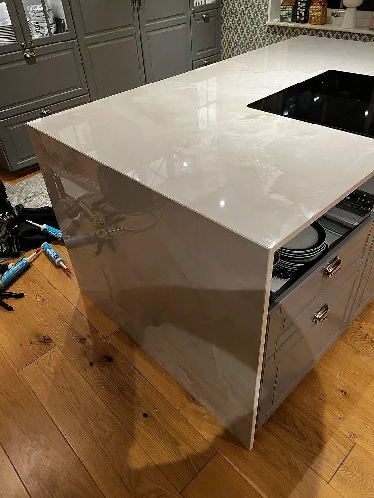 Sidovy av polerad Dekton Helena på köksö med neddragen stenpanel, svart häll och grågröna luckor i Växjö.