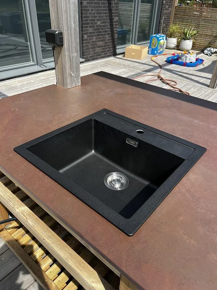 Svart diskho monterad under en matt Dekton Kadum bänkskiva i ett utekök på trädäck i Påarp.
