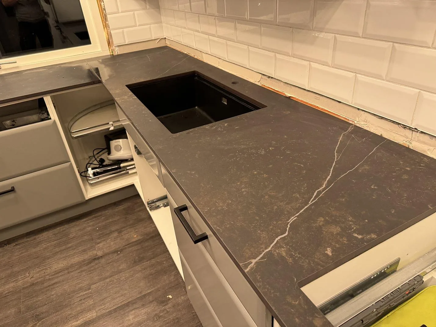 L-formad köksbänkskiva i matt Dekton Kelya med svart diskho, vit tunnelkakel och vita skåpsstommar i ett kök i Borås.