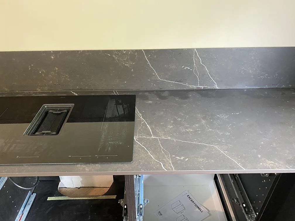 Köksbänkskiva i mörk Dekton Kelya, matt, med infälld induktionshäll och ljusådrigt stänkskydd i ett kök i Genarp.