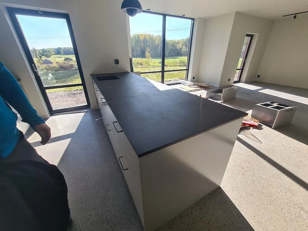 Köksö och lång köksbänkskiva i mörk matt Dekton Kelya keramik i ett kök med stora fönster i Glanshammar.