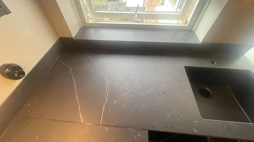 Matt köksbänkskiva i mörk Dekton Kelya med diskhoöppning, stänkkant och fönsterbänk i ett kök i Helsingborg.