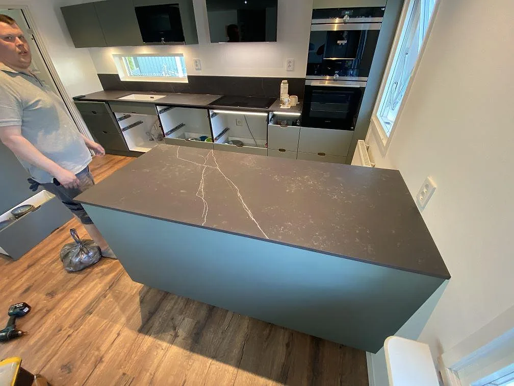 Köksbänkskiva i mörkgrå Dekton Kelya, matt, med köksö och längsgående bänk samt uppvikt stänkkant i ett kök i Höör.