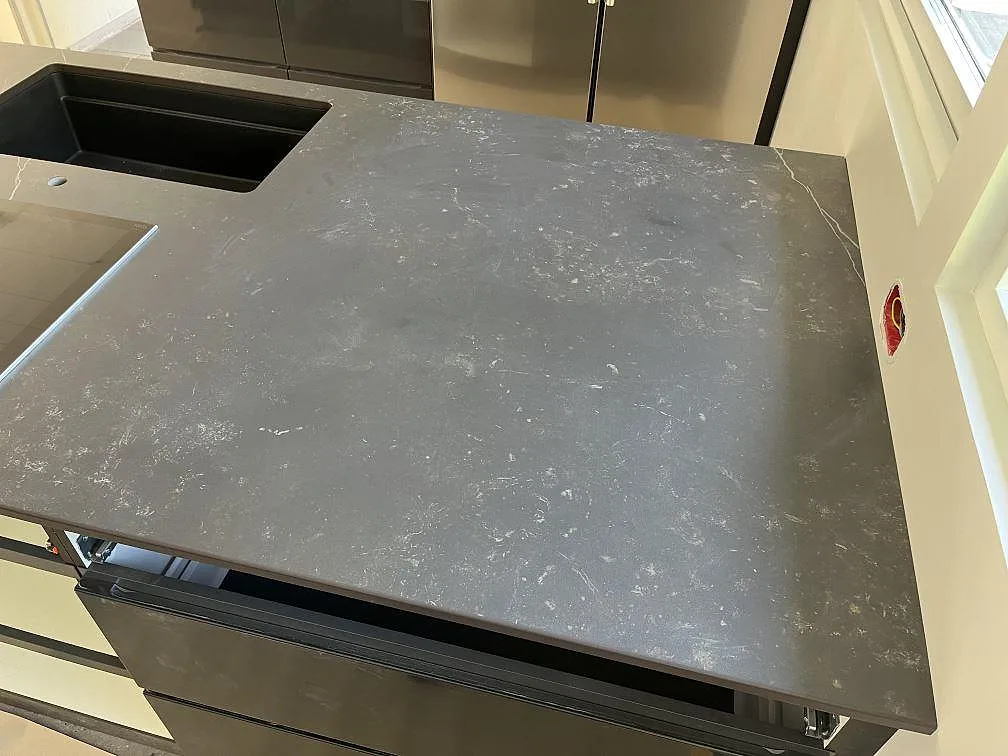 Köksö i grå Dekton Kelya, matt keramik med mjuk vit ådring, med svart underlimmad kompositdiskho i ett kök under montering i Löddeköpinge.