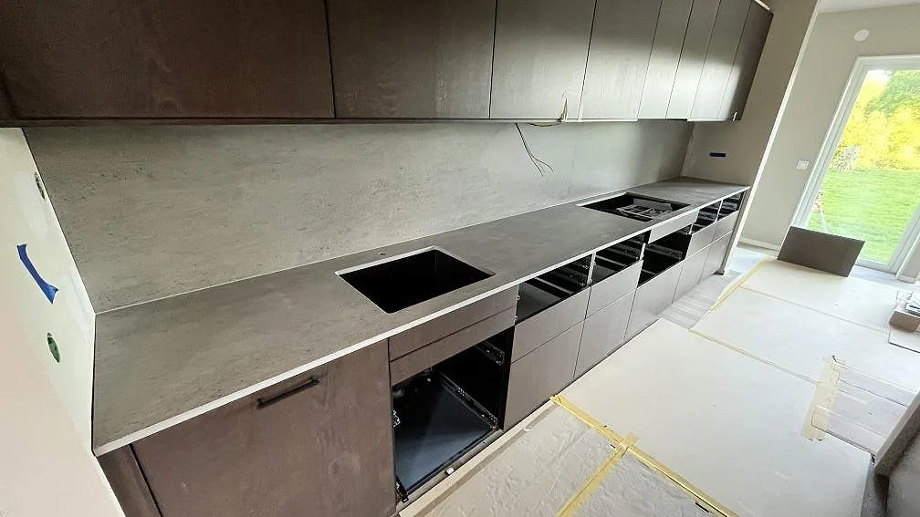 Lång köksbänkskiva i matt grå Dekton Keon med matchande väggskiva och urtag för diskho i ett kök under montering i Bestorp.